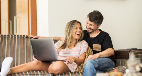 Un homme et une femme sont assis sur un canapé en riant, tandis que la femme tient un ordinateur portable sur ses genoux.